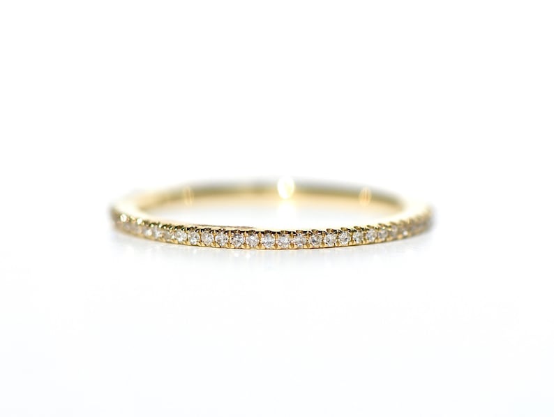 Puede incluir: Un delicado anillo de banda de oro, completamente engastado con peque&ntilde;os diamantes brillantes. El anillo se presenta sobre un fondo blanco brillante, resaltando los intrincados detalles y el brillo de las piedras preciosas.