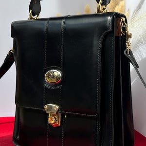 May include: Black leather briefcase with gold-coloured hardware. It has a top handle, a flap closure with a lock, and a detachable shoulder strap. The rectangular briefcase has a classic design, suitable for professional use.