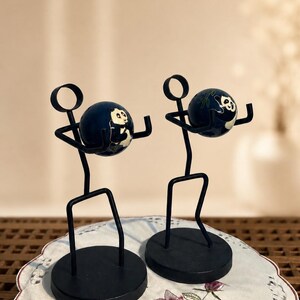 May include: Two black metal stick figure sculptures standing on circular bases. Each figure has a circular head and holds a dark blue spherical ornament featuring a white and black panda bear illustration with green bamboo accents.