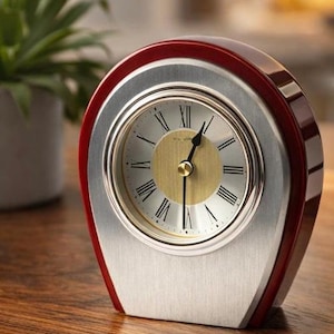 Peut inclure: Une horloge de table décorative avec un cadran argenté et des chiffres romains. L'horloge a un centre doré et des aiguilles noires. Le boîtier est une combinaison d'argent et de rouge foncé. L'horloge est sur une surface en bois.