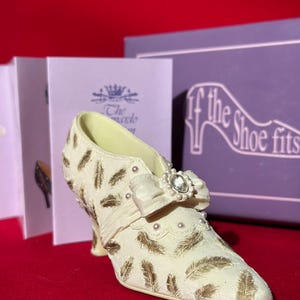 Puede incluir: Zapato decorativo de color marfil con detalles de plumas doradas, un lazo y una joya. El zapato está sobre una superficie roja, con una caja morada y un folleto en el fondo. La caja dice "If the Shoe Fits".