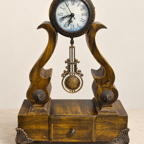 Relógio de mesa vintage ornamentado em tom dourado com pêndulo e gaveta, mostrador com algarismos romanos, detalhes de cabeça de leão, decoração de prateleira em estilo antigo.