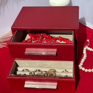 Op de afbeelding: Een rode sieradendoos met twee open lades. De bovenste lade bevat een rode koraal ketting en de onderste lade bevat diverse ringen en kettingen. Een parelketting is rechts zichtbaar.