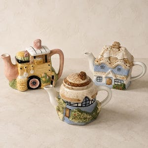 Peut inclure: Trois théières en céramique en forme de petits bâtiments. L'une est une maison jaune style chariot avec une anse rose, une autre est un cottage bleu au toit de chaume, et la troisième est un petit cottage bleu et blanc au toit brun texturé.