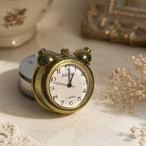 May include: A small, round, brass-toned alarm clock with a weathered finish. The clock face is off-white with black Arabic numerals, black hands, and the text SAIHO and 17 JEWELS printed in black. It features two bells on top and a winding knob.