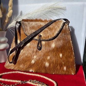 Könnte beinhalten: Eine rechteckige Handtasche mit braun-weiß geflecktem Fell außen. Die Tasche hat einen dunkelbraunen Lederriemen und einen dekorativen Verschluss. Eine Perlenkette liegt auf einer roten Oberfläche vor der Tasche.
