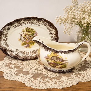 May include: A cream-coloured ceramic gravy boat and an oval platter, both featuring a dark brown floral border and a central illustration of game birds in a natural landscape. The pieces have a scalloped edge and a glossy finish.