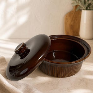 May include: A dark brown ceramic casserole dish with a lid. The dish has a ribbed exterior and a glossy finish. The lid is slightly ajar, revealing the interior. The dish is on a neutral surface.