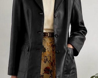 Könnte beinhalten: Ein schwarzer Lederblazer mit steigendem Revers, drei Knöpfen auf der Vorderseite und zwei aufgesetzten Taschen. Der Blazer wird über einem cremefarbenen Strickoberteil und einem plissierten Rock mit buntem Blumenmuster in Gelb, Rot und Grün getragen.