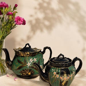 May include: Two ceramic teapots featuring a dark green and black color scheme with gold trim. The teapots are decorated with hand-painted floral motifs in white and pink, featuring scalloped edges on the lids and a glossy finish throughout.