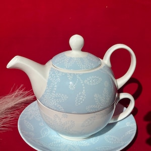 Könnte beinhalten: Ein weißes und hellblaues Teeservice mit floralem Muster. Das Set beinhaltet eine Teekanne, eine Tasse und eine Untertasse. Die Teekanne hat einen gebogenen Ausguss und Henkel. Die Tasse passt in die Teekanne, und die Untertasse befindet sich darunter. Das Set befindet sich auf rotem Hintergrund.