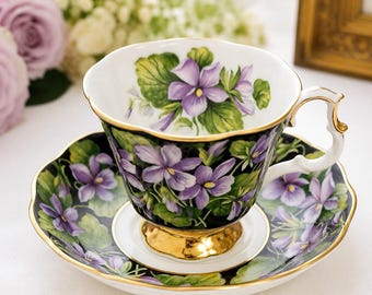 May include: A porcelain teacup and saucer set featuring a black background adorned with purple violet flowers and green leaves. The teacup has a gold-trimmed scalloped rim, a gold-painted pedestal base, and a white interior with floral accents.