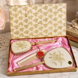 Puede incluir: Un set de tocador en una caja con estampado dorado y forro de satén rosa, que contiene un cepillo para el cabello, un peine y un espejo de mano. Cada pieza presenta un acabado blanco hueso con un diseño floral dorado y un mango dorado estriado.