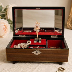 Può includere: Un portagioie rettangolare in legno con finitura scura e una placca per serratura color argento. L'interno è foderato in velluto rosso e presenta una ballerina rotante con tutù bianco, uno specchio sul coperchio e una piccola chiave in metallo.