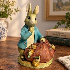 May include: A figurine of a rabbit wearing a blue jacket and holding a large brown sack with blue polka dots. The rabbit has light brown fur, long ears, and is standing on a textured base. The sack contains visible green leafy vegetables.