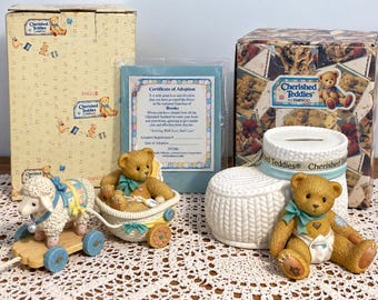 Puede incluir: Una colección Cherished Teddies que incluye una figura de cordero en un juguete de arrastre de madera, un oso de peluche en un cochecito y un oso de peluche junto a una hucha de cerámica blanca con forma de zapato, con dos cajas de cartón.
