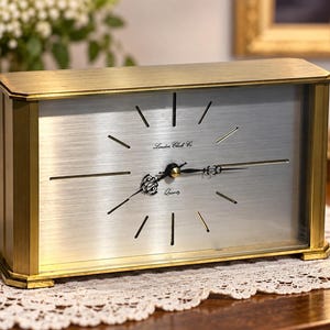 May include: A rectangular mantel clock with a brushed silver face and a gold-toned metal frame. The clock features black hands, thin black hour markers, and the text London Clock Co and Quartz printed in black on the centre of the dial.