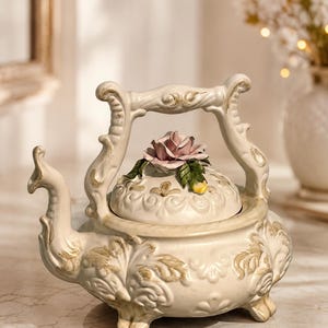 Peut inclure: Une théière en céramique couleur crème avec une anse ornée, un bec verseur incurvé et trois petits pieds. Le couvercle est décoré d'une rose rose sculptée et de feuilles vertes, tandis que le corps présente des motifs floraux en relief dorés.