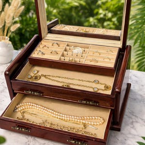 May include: A dark wood jewelry box with a mirrored lid, open to reveal multiple compartments. The top tray holds rings and small jewelry, while the drawers below contain necklaces and bracelets. The box is filled with gold and pearl jewelry.