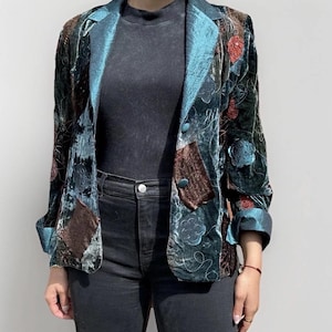 May include: A teal velvet blazer with a teal satin lapel and cuffs. The blazer features embroidered floral details, brown patches, and teal buttons. The sleeves are cuffed, revealing the satin lining. The blazer is worn over a black top and black jeans.