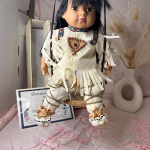 Peut inclure: Poupée en porcelaine représentant un enfant amérindien, suspendue par des cordes. La poupée porte une tenue à franges crème avec des perles et des mocassins. La poupée a des cheveux noirs et est assise sur une balançoire. Un certificat est visible.