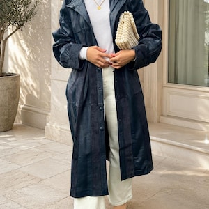 May include: A dark navy blue leather trench coat worn over a white ribbed turtleneck sweater and white wide-leg trousers. The outfit includes a cream-colored beaded handbag and pointed-toe shoes with a metallic, textured finish.