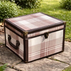 May include: A plaid pattern storage trunk with brown leather trim and brass corner protectors. The trunk has a handle and a latch closure. The trunk is sitting on a stone surface, with greenery in the background.