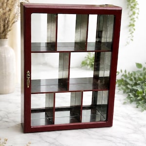 May include: A burgundy display cabinet with a glass door and multiple shelves. The cabinet has a rectangular shape with a brass-coloured handle and hinges. The shelves are divided into various compartments.