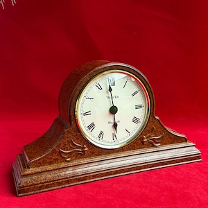 May include: A brown wooden mantel clock with a cream-colored face and gold trim. The clock face features black Roman numerals and the words "Westclox Warwick". The clock has a curved top and decorative carvings.
