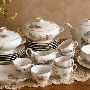 Może przedstawiać: Zestaw białej porcelany z delikatnym botanicznym wzorem brązowych i szarych liści ze złotym wykończeniem. Kolekcja zawiera misy do serwowania z pokrywkami, dzbanek do herbaty, mlecznik, kilka filiżanek, spodki oraz owalny półmisek do serwowania.