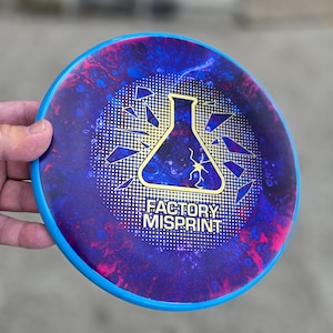 May include: A blue and purple disc golf disc with a blue rim. The disc features a gold beaker graphic with a lightning bolt and the text "FACTORY MISPRINT". The disc has a marbled design with pink and blue accents.