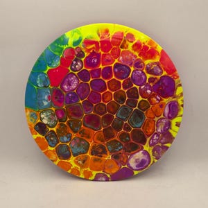 May include: A circular abstract artwork with a vivid, cell-like pattern. The piece showcases a range of colours, including yellow, red, orange, purple, and blue. The design resembles a close-up view of cells or a honeycomb structure.