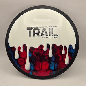 May include: A white and black disc golf disc with the word "TRAIL" in black, and the text "JAMES CONRAD | 2021 WORLD CHAMPION". The disc has a white, dripped paint design over a red, blue, and black pattern.