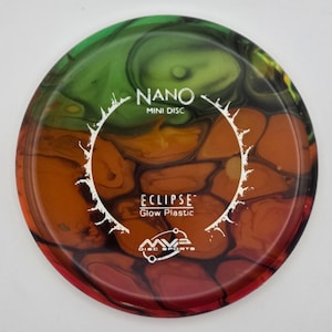 Puede incluir: Un mini disco de golf con un diseño veteado en tonos verdes, naranjas y rojos. El disco presenta el texto "NANO MINI DISC" y "ECLIPSE Glow Plastic" en blanco. También es visible el logotipo de MVA Disc Sports.
