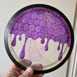 May include: A clear disc golf disc with a black rim. The disc features a purple honeycomb pattern and dripping purple paint. The center has a graphic of a broken beaker with the text "Lab Second".