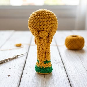 Könnte beinhalten: Handgefertigte Häkeltrophäe in Gelb- und Grüntönen. Die Trophäe hat einen runden Kopf, einen detaillierten Körper und einen grün-gelben Sockel. Ein Knäuel gelbes Garn und eine Häkelnadel sind ebenfalls zu sehen, was auf ein Bastelprojekt hindeutet.