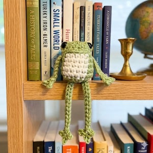 Puede incluir: Un marcapáginas de rana de ganchillo hecho a mano en tonos verdes y blancos, con ojos negros, está sobre una estantería de madera. Las largas patas de la rana cuelgan sobre los libros. Se ven libros con títulos como "A History of Tales".