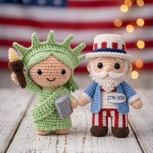 May include: Crocheted figures of the Statue of Liberty and Uncle Sam. The Statue of Liberty is green, holding a torch and tablet. Uncle Sam wears a blue coat, striped pants, and a hat. The figures are set against an American flag.