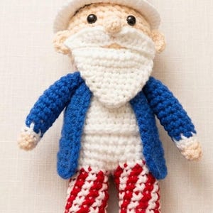 May include: A handmade crochet doll depicting Uncle Sam. The doll wears a white hat with red and blue stripes, a white beard, a blue jacket, and red and white striped pants. A silver coin is attached to the hat.