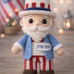 Op de afbeelding: Een gehaakte Uncle Sam-pop, gekleed in een blauw jasje, rood-wit gestreepte broek en een hoed met rood-blauwe strepen. De pop heeft een witte baard en een label met de tekst "1776-2026".