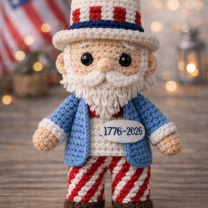 May include: A crocheted Uncle Sam doll with a white beard, blue jacket, and red and white striped pants. The doll wears a white hat with red and blue stripes. The text "1776-2026" is on the doll's chest. An American flag is in the background.