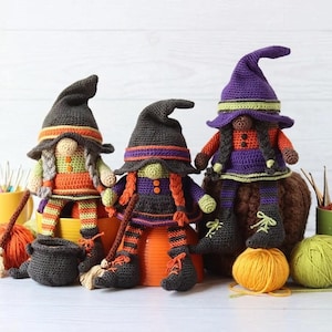 May include: Three crocheted dolls dressed as witches, featuring pointed hats in dark grey and purple, orange and black striped leggings, and small broom accessories. The dolls are displayed with yarn balls, crochet hooks in mugs, and a small black cauldron.