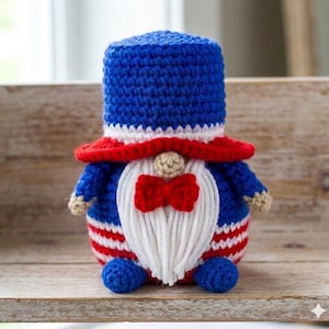 Può includere: Una bambola gnomo all'uncinetto patriottica con un cappello a cilindro blu, un corpo a righe rosse e bianche e una barba bianca con un papillon rosso. Lo gnomo è seduto su una superficie di legno.