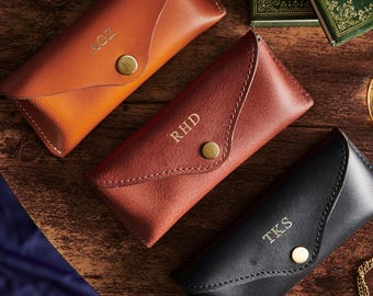 May include: Three leather eyeglass cases in shades of brown and black, each with a personalized monogram. The cases have a flap closure with a gold-colored button. The cases are on a wooden surface.