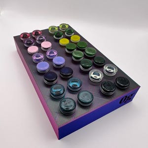 Puede incluir: Una exhibición de coloridos tapones de vidrio dispuestos sobre una plataforma rectangular morada y azul. Los tapones son de varios colores, incluyendo rosa, verde, azul y negro. La plataforma tiene una superficie texturizada y un número impreso en el lateral.