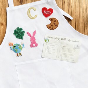 Puede incluir: Un delantal blanco con varios parches bordados que incluyen una letra C amarilla, un corazón rojo con XOXO, una galleta con chispas de chocolate, un trébol verde, un conejo rosa y una Tierra de dibujos animados con un cartel. Incluye una tarjeta de receta Earth Day Jello Aquarium.