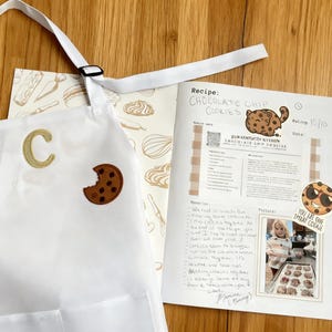 May include: A white apron featuring a gold embroidered letter C and a brown chocolate chip cookie patch. Beside it lies a recipe sheet titled Chocolate Chip Cookies with a cartoon cookie illustration, a QR code, and a handwritten note.