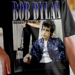 May include: Black t-shirt featuring a graphic of Bob Dylan with the text "BOB DYLAN HIGHWAY 61 REVISITED." The image shows a vintage photo of Bob Dylan wearing a patterned jacket and a white t-shirt.