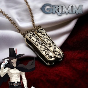 Puede incluir: Un collar con colgante dorado con dos amuletos rectangulares con intrincados diseños grabados. El collar tiene una cadena delicada y la palabra "GRIMM" es visible en el fondo. La estética general sugiere un tema de fantasía o gótico.