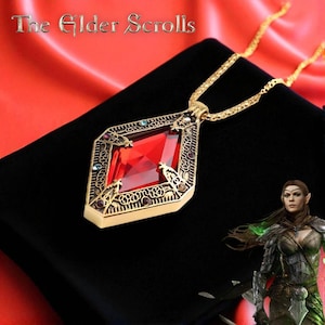 Puede incluir: Collar con colgante dorado con una gran gema roja facetada. El colgante tiene un diseño ornamental de filigrana con pequeñas piedras de acento de colores. La cadena también es dorada. El texto "The Elder Scrolls" está en la parte superior de la imagen.